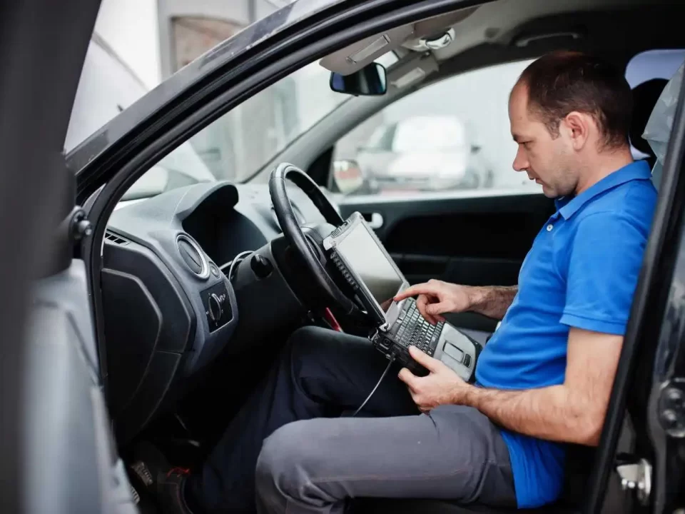 Imagem com um homem sentado no banco do carro, com um notebook em sua mão parecendo estar fazendo algum ajuste por meio dele.