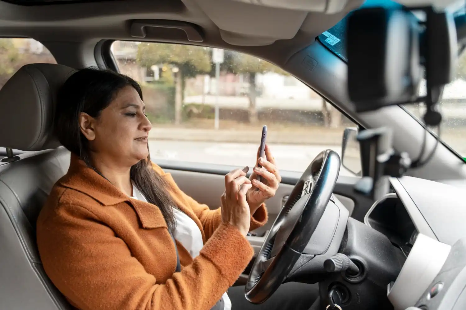 Mulher sentada no banco do motorista de um carro, usando o celular com as duas mãos, usando casaco laranja e cinto de segurança.