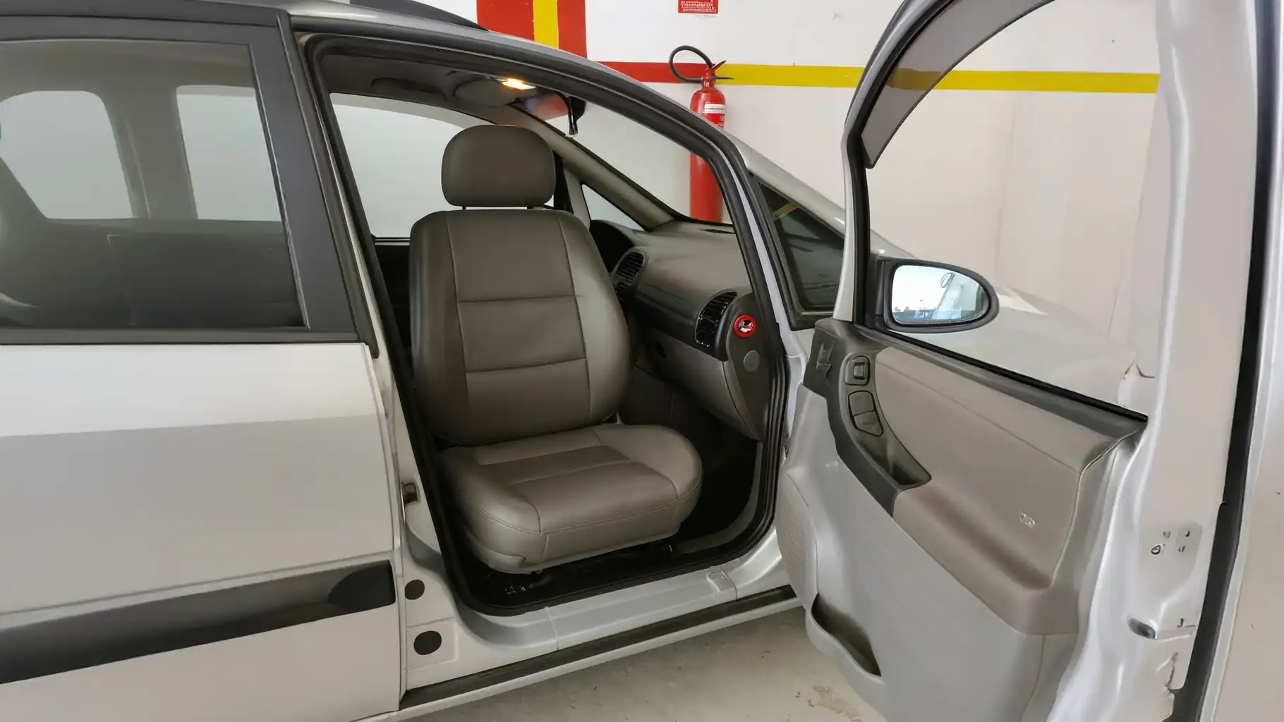 Interior de um carro prata com a porta do motorista aberta, mostrando um banco dianteiro de couro cinza claro girado para o lado da porta, com painel, volante e retrovisor visíveis.